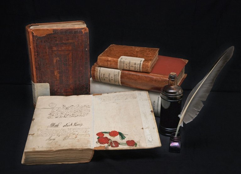 Foto av gamle bøker, den ene ligger oppslått på bordet. Fjærpenn i blekkhus ved siden av.