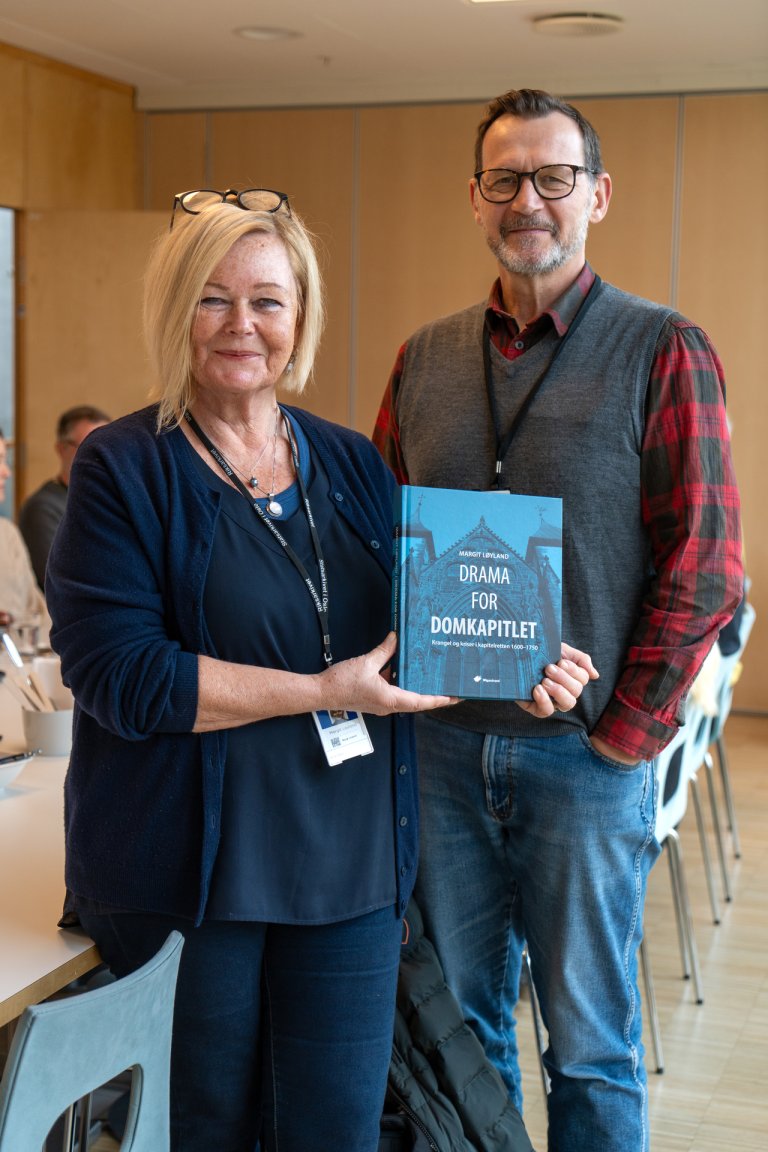 To smilende personer som holder en blå bok med tittelen Drama for Domkapitlet på et kontor med andre synlige bak seg.