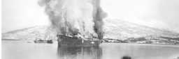 Det tidligere hurtigruteskipet "Dronning Maud" har blitt bombet av tyske fly. Skipet står i brann og mørk røyk velter opp mot himmelen.